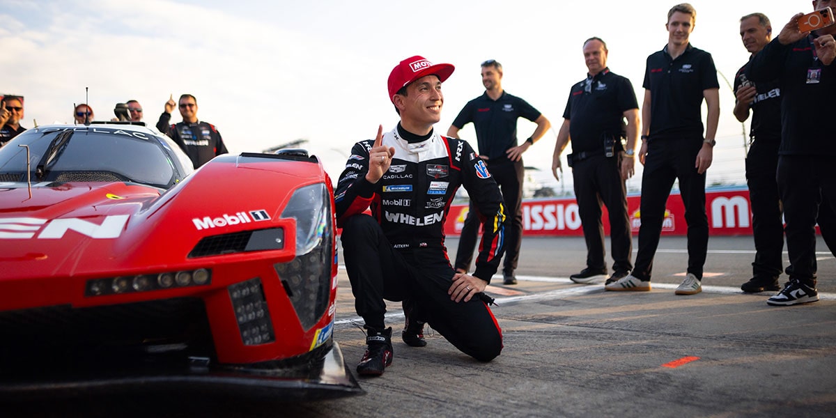 A Cadillac Racing Driver in a Black Whelen-Branded Suit Kneeling Beside a Red Cadillac V-Series.R Race Car on Pit Lane With Team Members and Equipment in the Background.