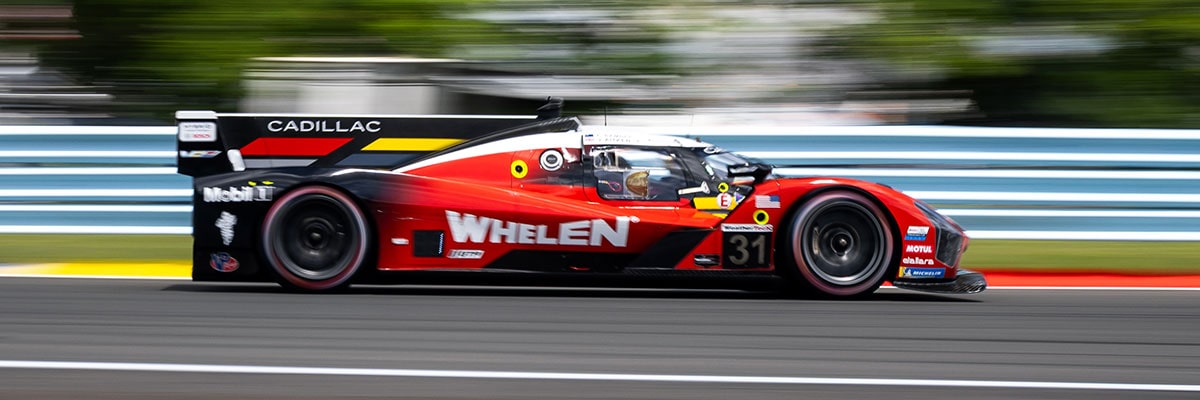 A Red Cadillac V-Series.R Race Car With Whelen Branding Driving at High Speed Along a Track With Motion Blur and Blue Guardrails Lining the Circuit.