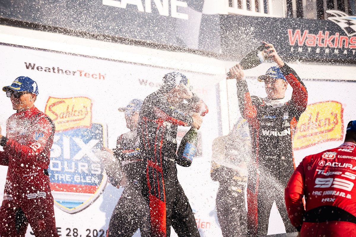Cadillac Racing Team Drivers Spraying Champagne on a Podium at Watkins Glen International With WeatherTech and IMSA Branding Visible on the Celebration Backdrop.