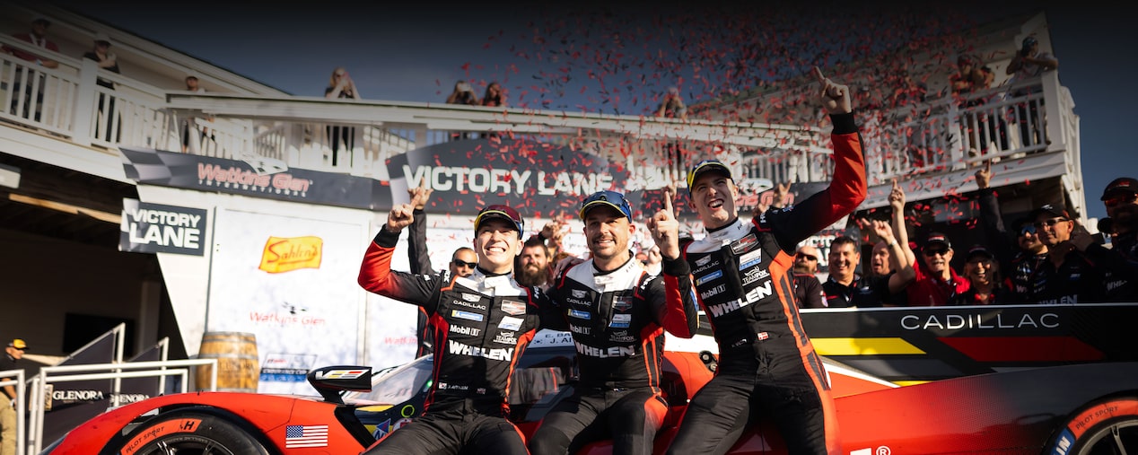 Three Cadillac Racing Team Drivers in Black Whelen-Branded Performance Suits Celebrating on a Red Cadillac Race Car in Front of a Victory Lane Sign With Red Confetti Falling Overhead.