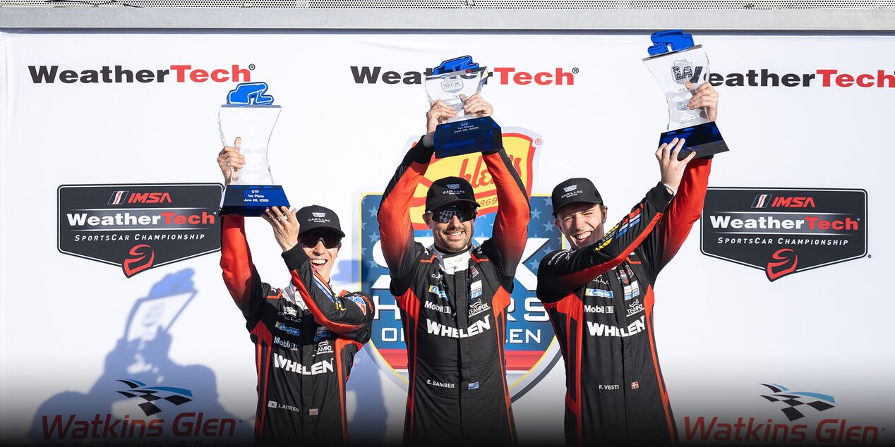 Three Cadillac Racing Team Drivers in Black Whelen-Branded Racing Suits Standing on a Podium at Watkins Glen International Holding WeatherTech and IMSA Trophies in Front of a Branded Backdrop Featuring Event Logos.