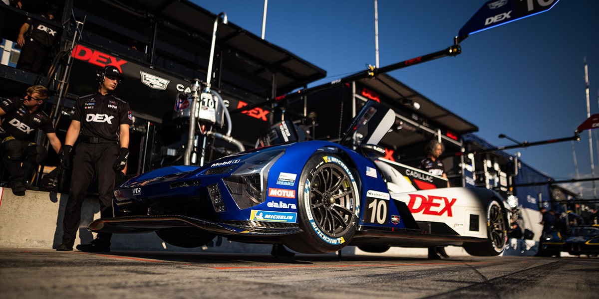 Cadillac V-Series.R Race Car Stopped on Track With Team Garage Structures Visible in the Background.