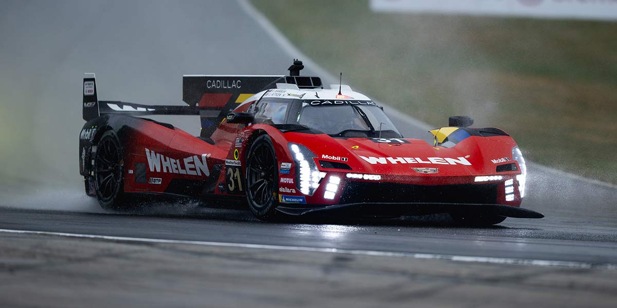 Un piloto de Cadillac Racing con un traje negro con la marca Whelen arrodillado junto a un auto de carreras Cadillac V-Series.R rojo en la calle de boxes con miembros del equipo y equipamiento en el fondo.