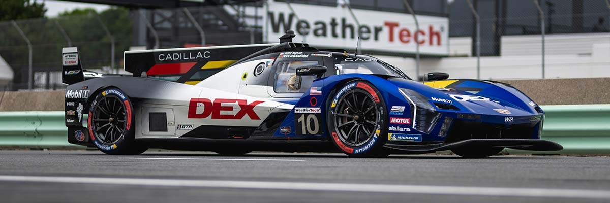 Un auto de carreras Cadillac V-Series.R rojo con la marca Whelen circulando a gran velocidad por una pista con efecto de desenfoque por movimiento y barandillas azules delimitando el circuito.