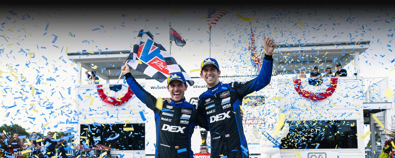 Tres pilotos del equipo Cadillac Racing, vestidos con trajes de competición negros con la marca Whelen, celebrando sobre un auto de carreras Cadillac Rojo frente a un letrero de Victory Lane, mientras cae papel picado rojo desde arriba.