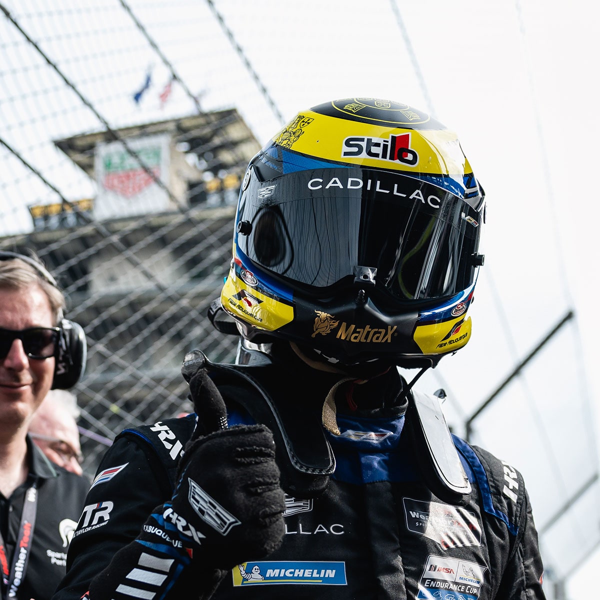 A Cadillac Racing Driver in a Racing Suit Raises Both Arms While Holding a Checkered Flag in Front of the Pit Area at a Racetrack.