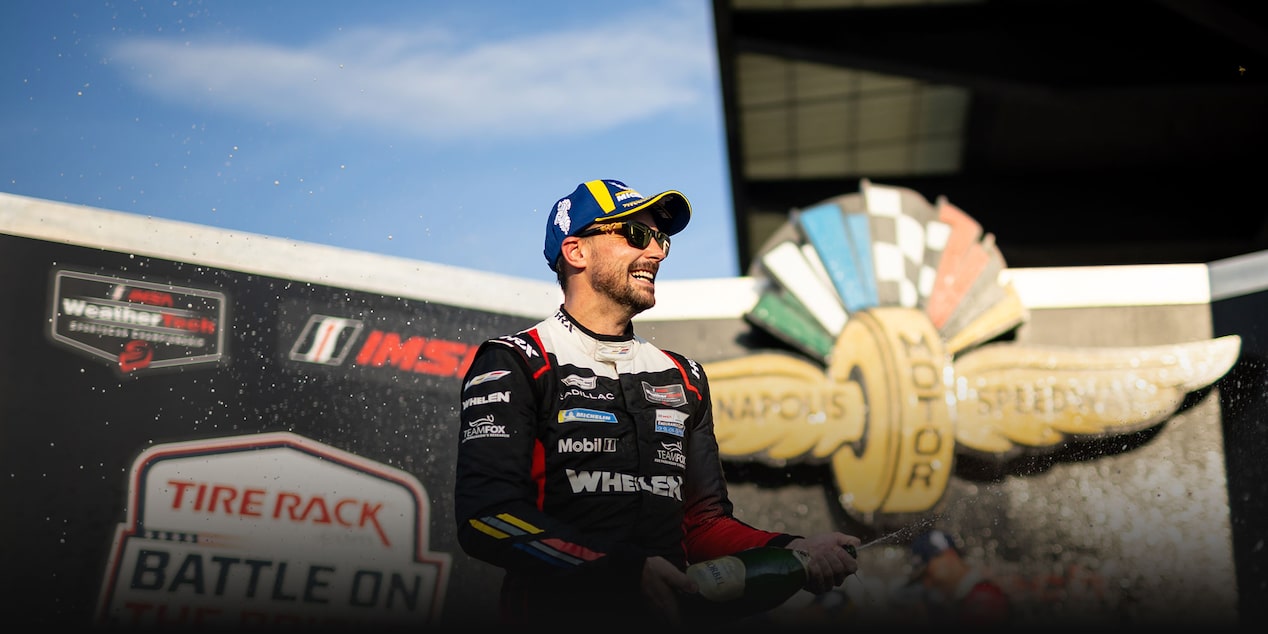 A Close-Up of Two DEX Racing Team Drivers in Blue and Black Racing Suits and Caps Raising Their Index Fingers Beside a Race Car Tire in Front of an IMSA SportsCar Endurance Championship Backdrop.