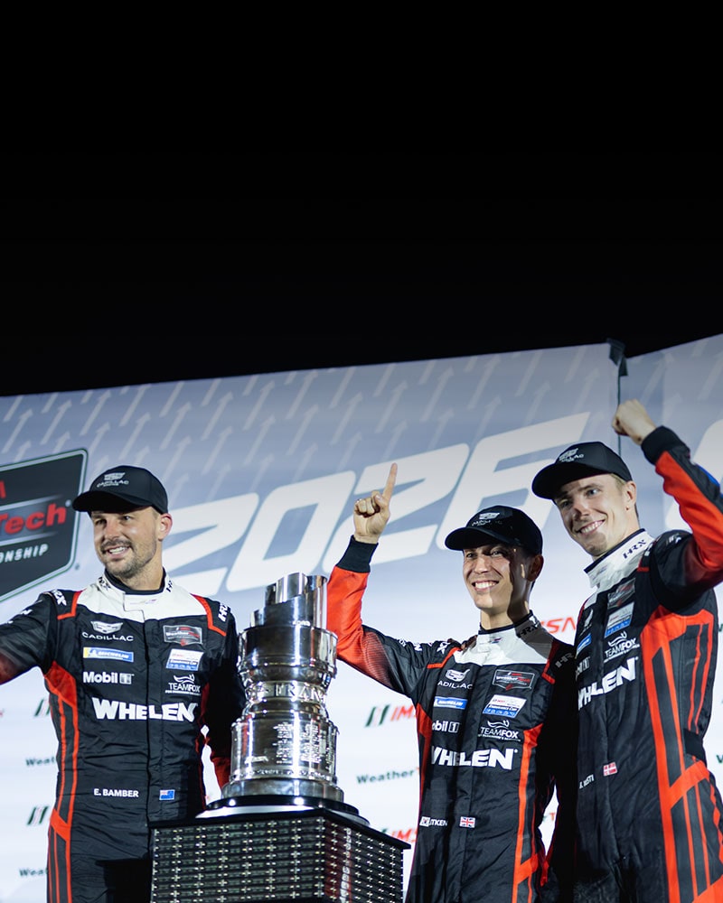 A Cadillac Racing Driver Standing in Front of a IMSA Backdrop Smiling and Holding a Champagne Bottle