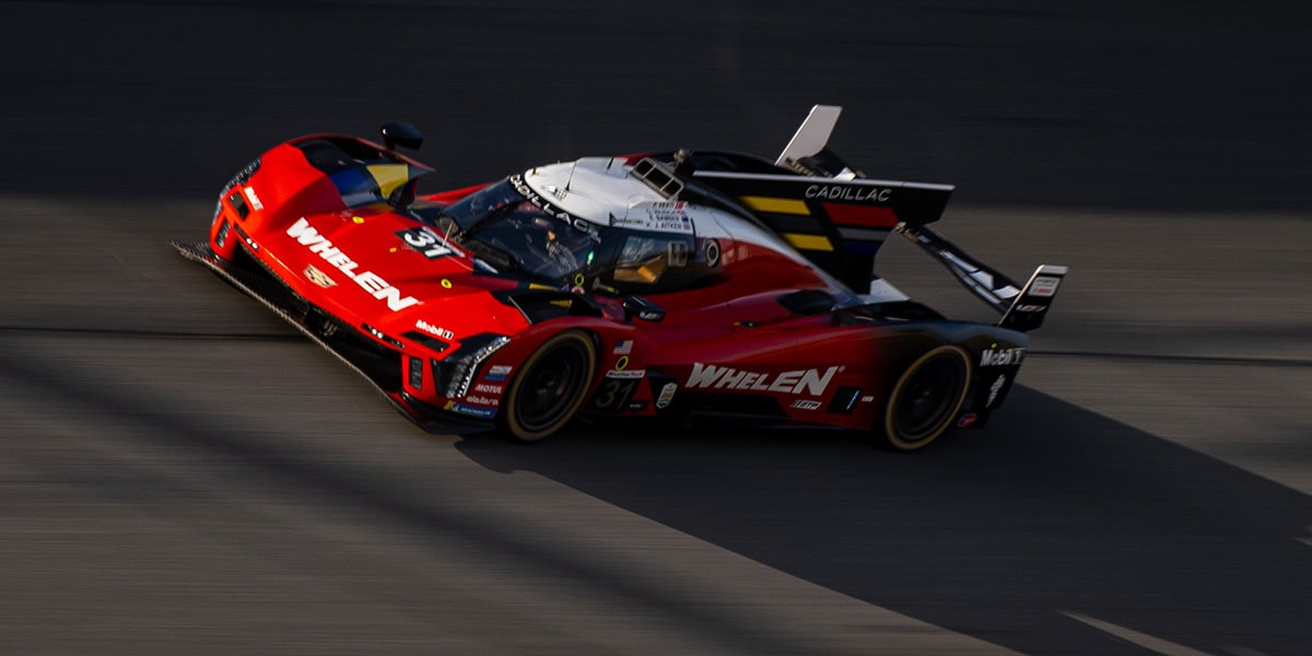 A Red Whelen Cadillac V-Series.R Race Car Races Down the Track at Dusk With Its Aerodynamic Body Low and Fast Against the Asphalt.