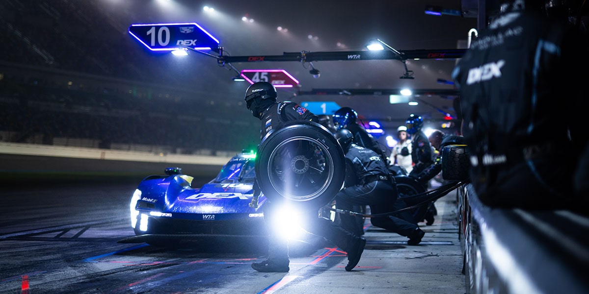 A Whelen Cadillac V-Series.R Race Car Enters the Pit Lane at Night With Intense Lights Illuminating the Front of the Vehicle and Crew Members Ready for Service.