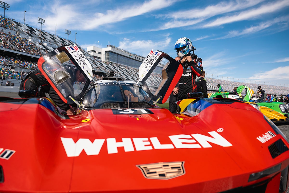 A Crew Member Works on a Red Whelen Cadillac V-Series.R Race Car in the Pit Lane Under Bright Daylight With the Vehicle's Hood Open and Tools Visible Around the Cockpit.