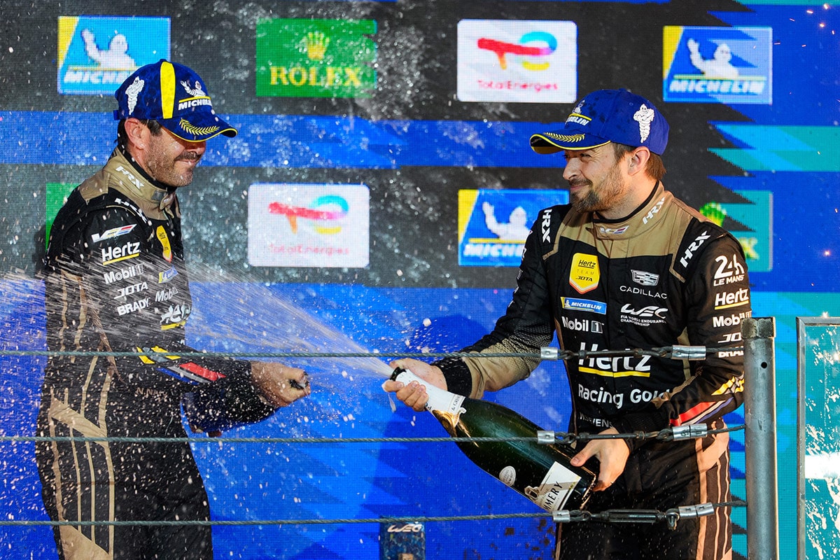 Drivers in Cadillac Hertz Team JOTA Racing Suits Spraying Champagne on a Podium During a Post-Race Celebration With Sponsor Logos Displayed in the Background.