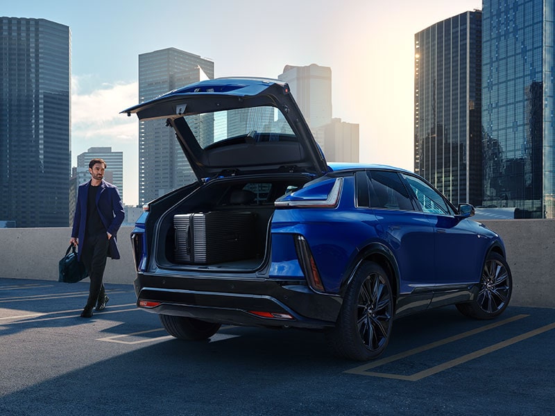 A Business Man Walking Around a Blue Cadillac LYRIQ Parked on a City Rooftop Parking Deck With the Power Liftgate Open