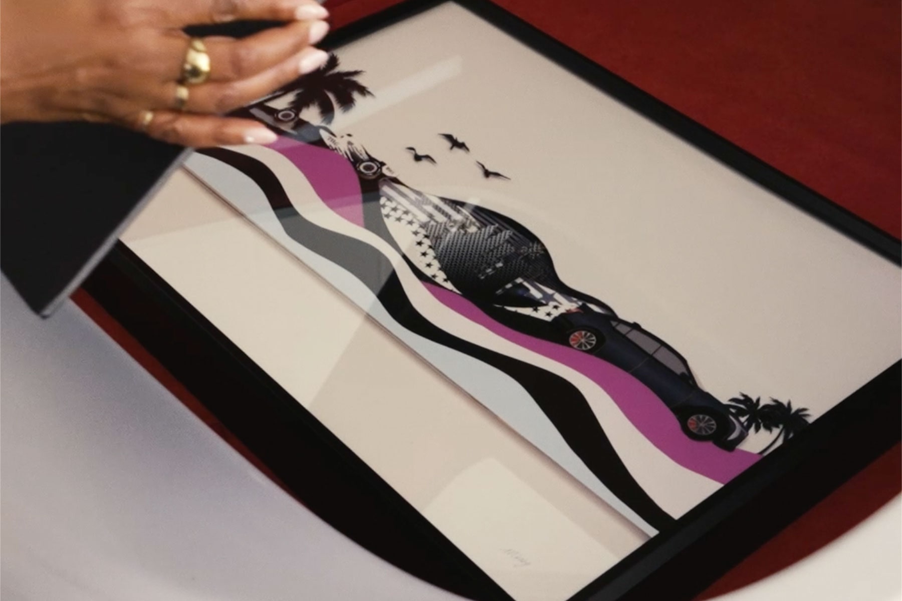 A Hand Opening a Framed Cadillac-Inspired Artwork Featuring a Vehicle Illustration, Checkered Flag, Palm Tree, and Flowing Abstract Design Elements on a Red Surface