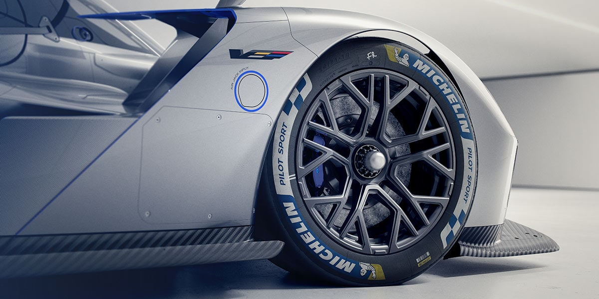 A Close-Up View of the Front Wheel and Side Bodywork of the Silver Cadillac V-ONE Concept Vehicle Featuring a Black Multi-Spoke Wheel, Red Brake Caliper, Michelin Tire Branding, and Carbon Fiber Aerodynamic Components.