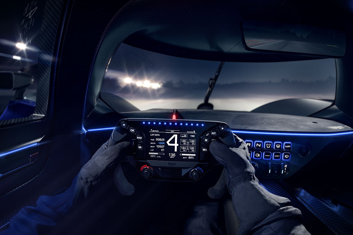 A Driver’s-Eye View From Inside the Silver Cadillac V-ONE Concept Vehicle Showing Gloved Hands on a Digital Steering Wheel, an Illuminated Instrument Display, Blue Ambient Interior Lighting, and a Race Track Visible Through the Windshield at Night.