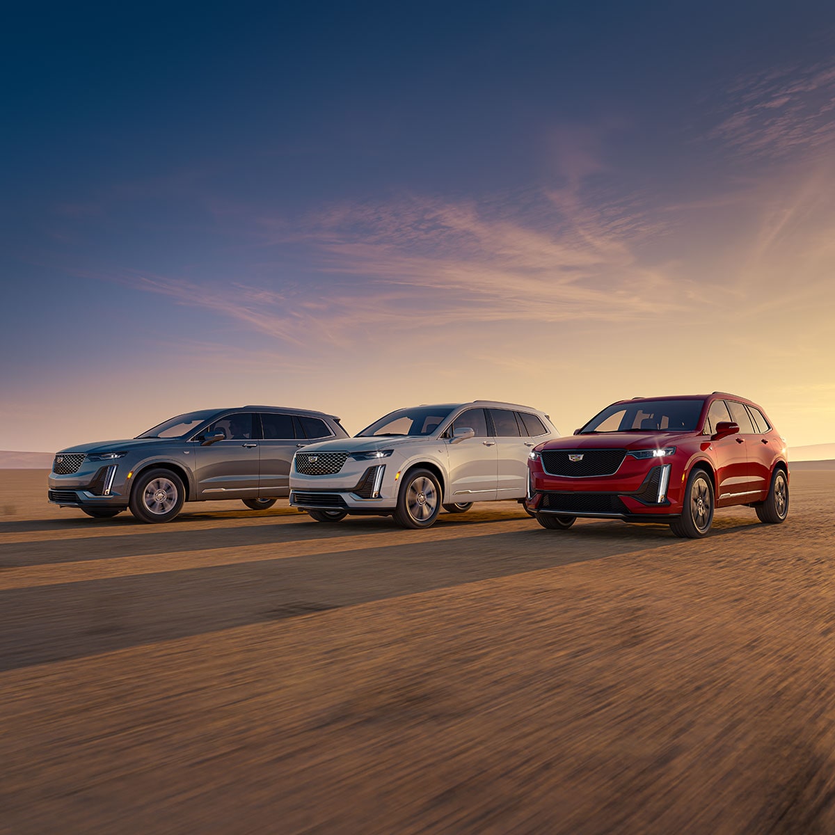 Vista de medio perfil de tres Cadillac XT6 en Plata, Blanco y Rojo estacionadas, con un atardecer a lo lejos