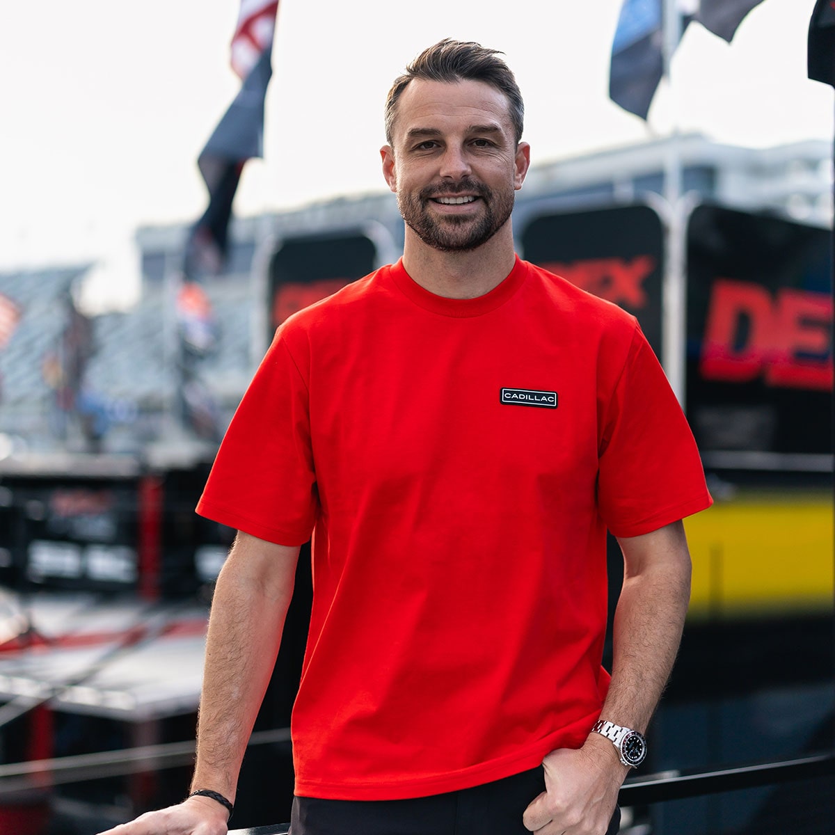 A Man Wearing a Red T-Shirt With a Small Rectangular Logo Stands in Front of Outdoor Event Equipment.