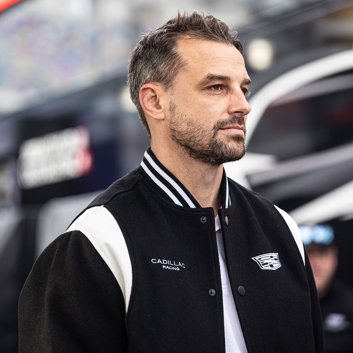 A Man Wearing a Black and White Jacket With Cadillac Racing Logos Stands in Front of Equipment at a Motorsports Event.
