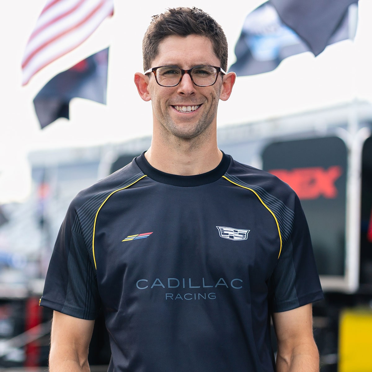 A Man with Short Brown Hair Wearing a Black Cadillac Racing Shirt Stands Outdoors in Front of Racing Team Flags and Equipment at a Motorsport Event.