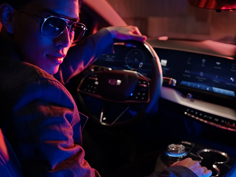 Conductor con gafas de sol mirando detrás del hombro, con la mano en el volante de una Cadillac OPTIQ 2026