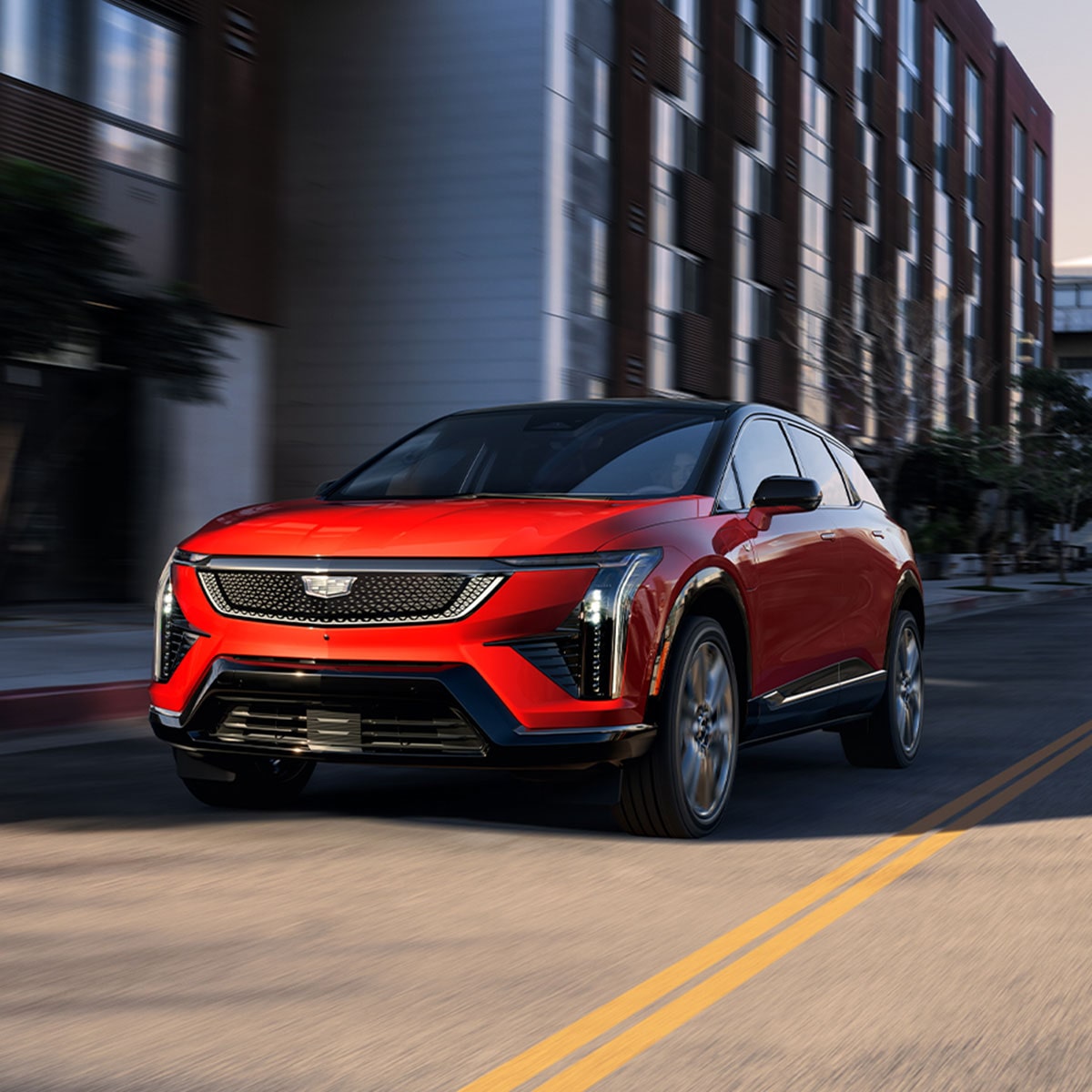 A Red 2026 Cadillac OPTIQ Driving Down a City Street with Buildings in the Background