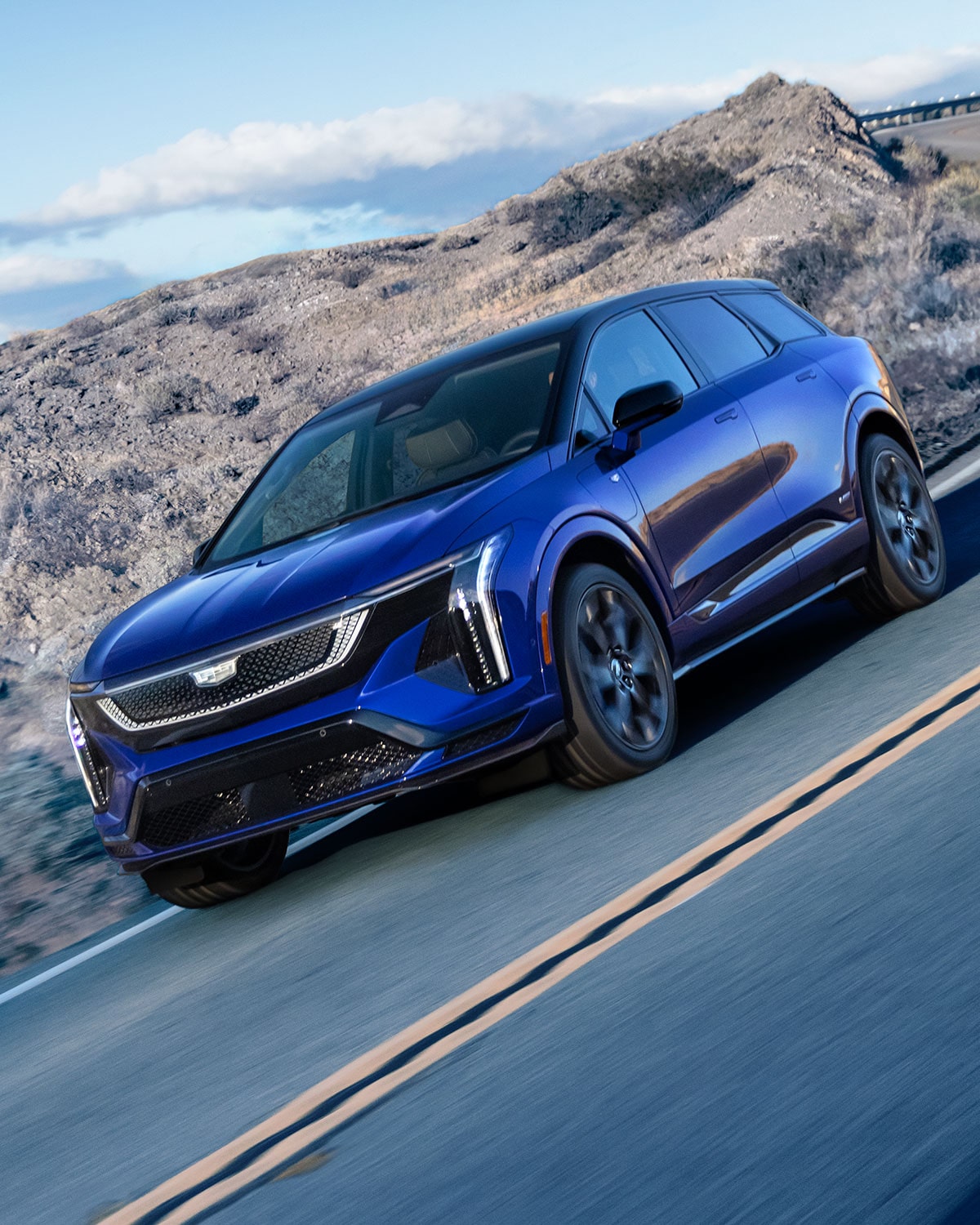 A Blue Cadillac OPTIQ-V Driving Around the Curve of a Mountain Road