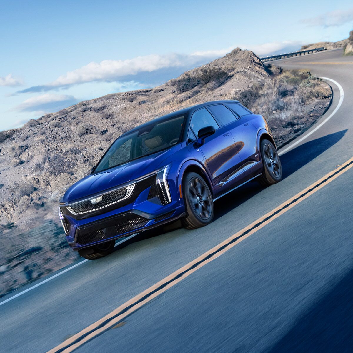 A Blue Cadillac OPTIQ-V Driving Around the Curve of a Mountain Road