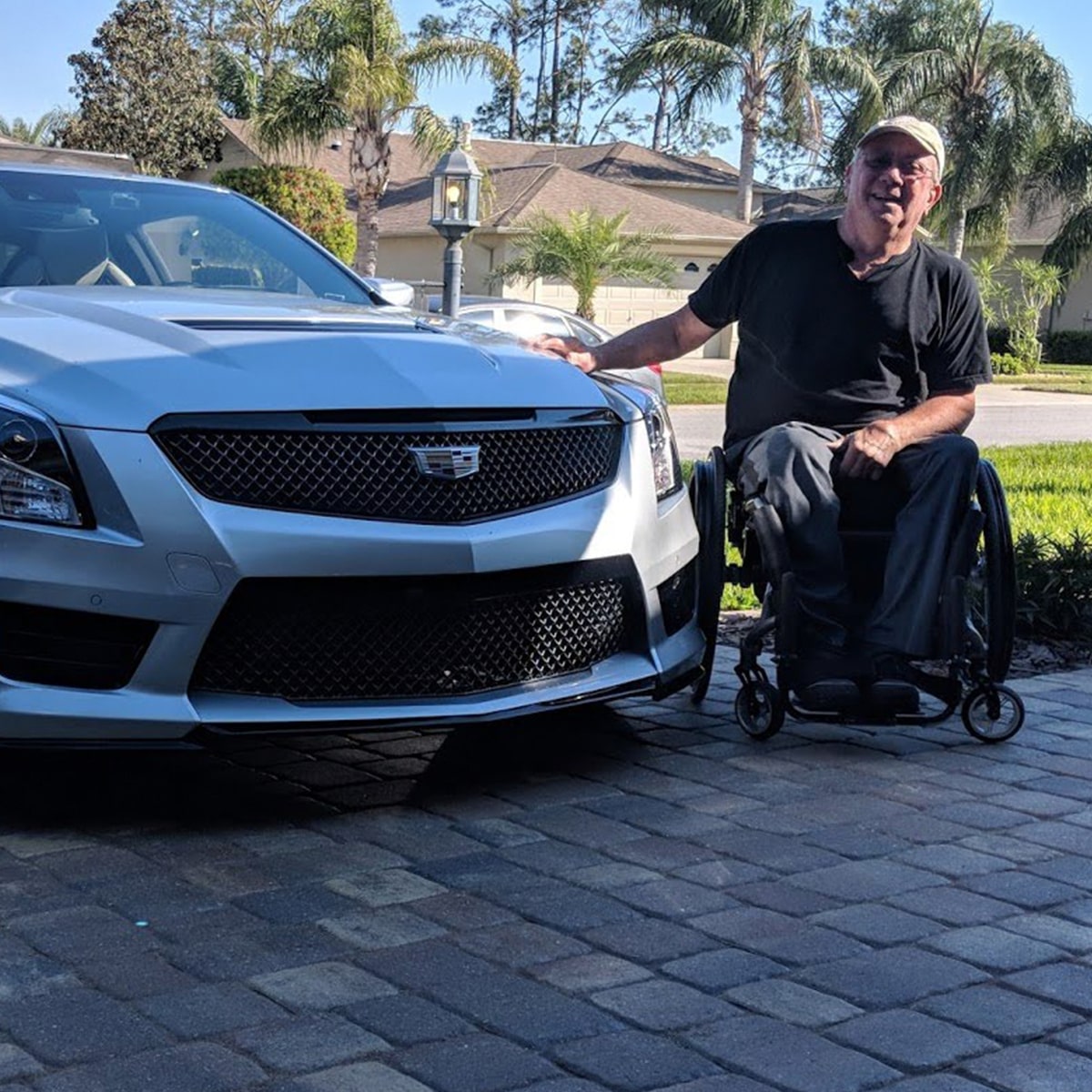 Hombre en silla de ruedas sonriendo, con una mano en el cofre de un sedán Cadillac V-Series, y una casa y palmeras de fondo