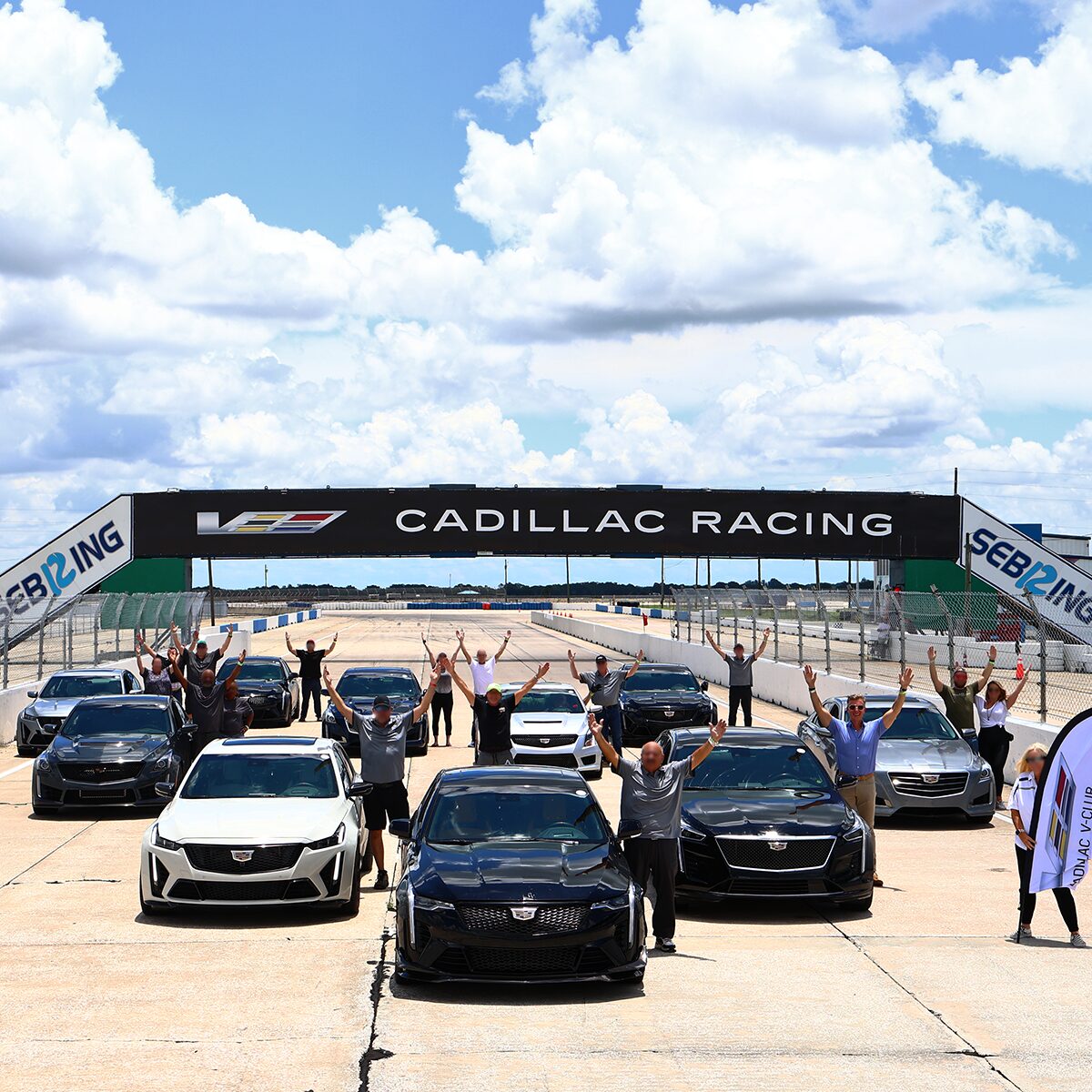Sedanes Cadillac V-Series estacionados en filas, debajo del cartel de Cadillac Racing en una pista de carreras