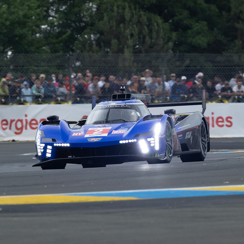 Blue Cadillac V-Series.R Race Car With Head Lights On On The Track And Crowd In The Background