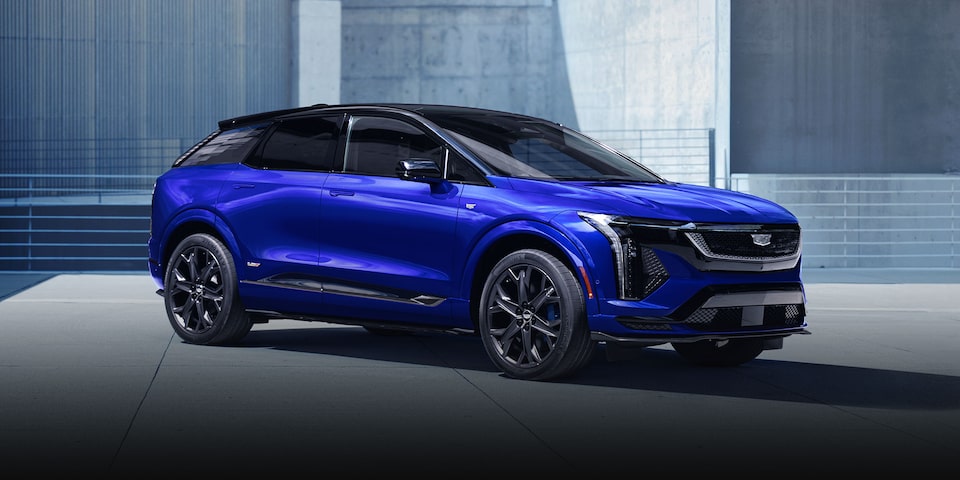A Side View of a Blue Cadillac OPTIQ-V with a Black Roof and Dark Alloy Wheels Parked in a Clean Urban Setting with Modern Architectural Walls in the Background.
