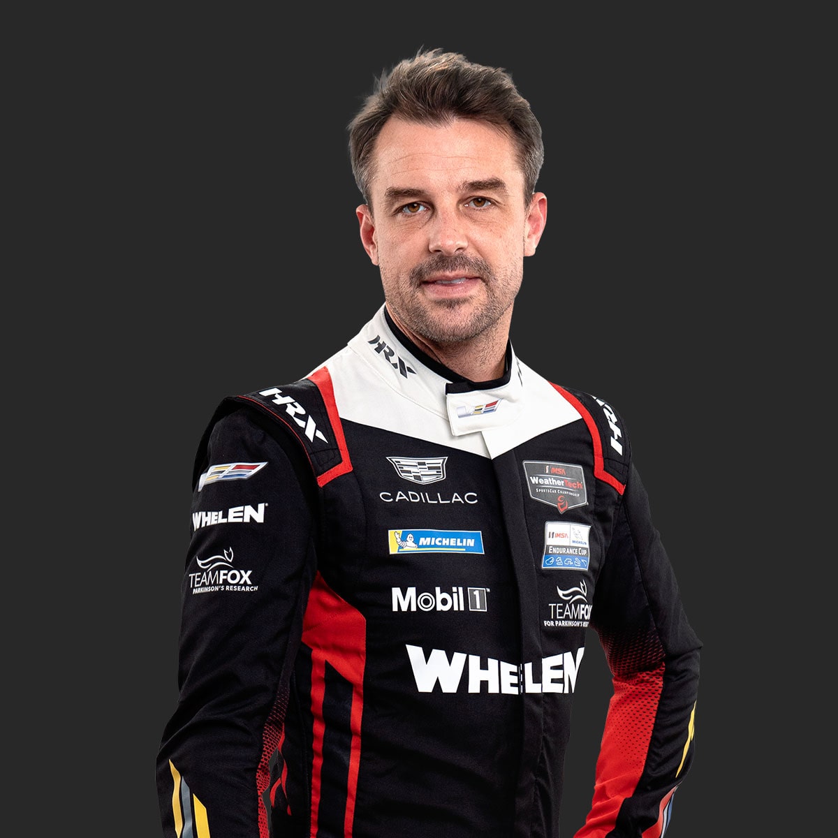 Cadillac Racing Team Driver Earl Bamber Wearing a Black, White, and Red Whelen‑Branded Racing Suit With Sponsor Patches Stands With Both Arms at the Sides Against a Solid Dark Background.