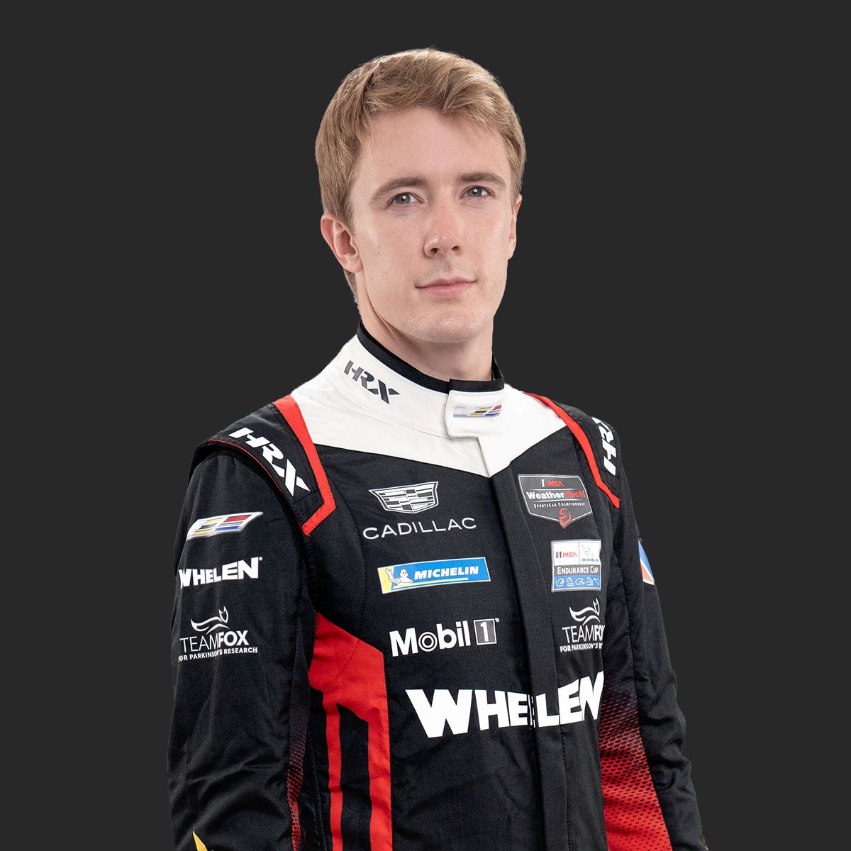 Cadillac Racing Team Driver Frederik Vesti Wearing a Black, White, and Red Whelen‑Branded Racing Suit With Sponsor Patches Stands Against a Solid Dark Background.