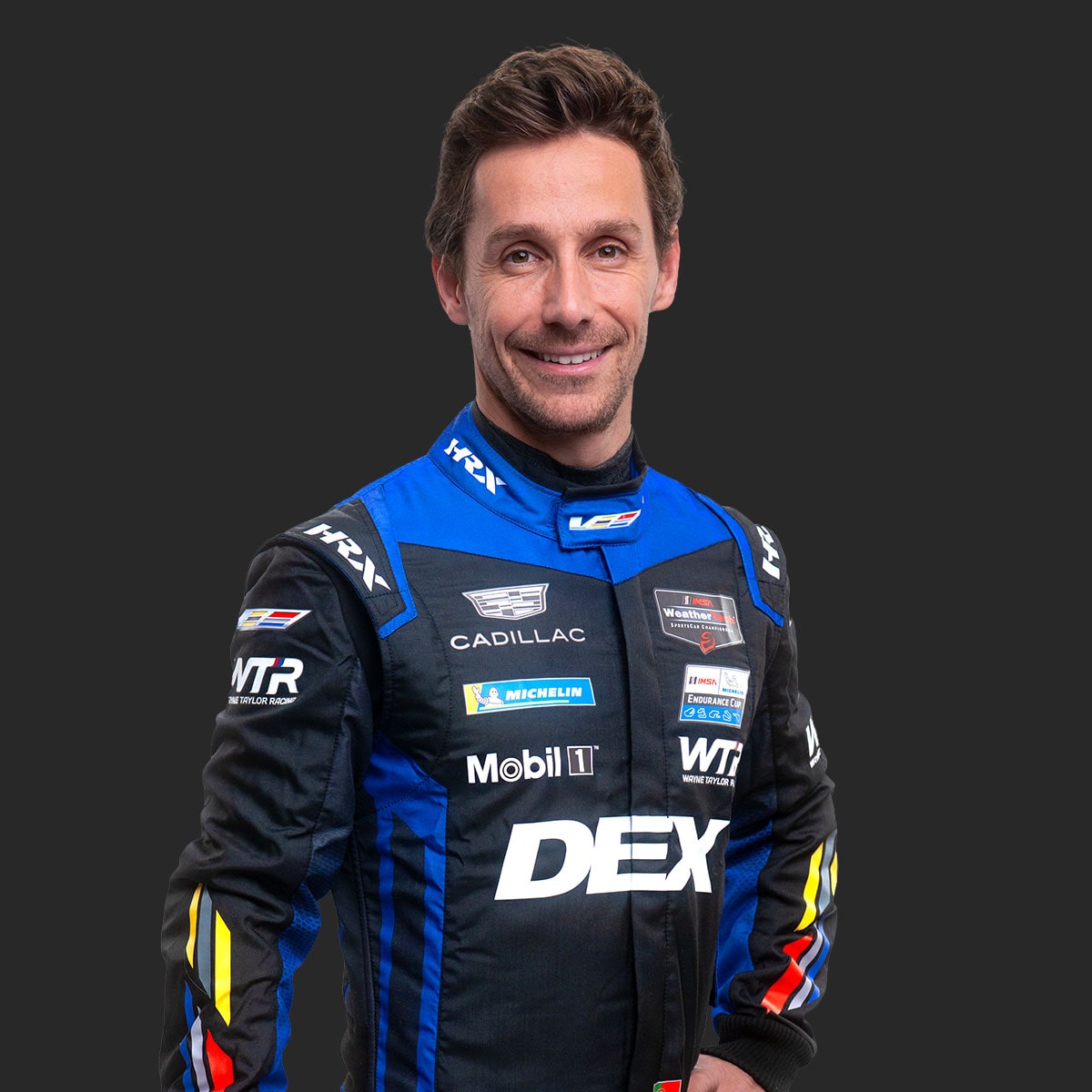 Cadillac Racing Team Driver Filipe Albuquerque Wearing a Blue and Black DEX Imaging–Branded Racing Suit Is Shown Standing Against a Solid Dark Background.