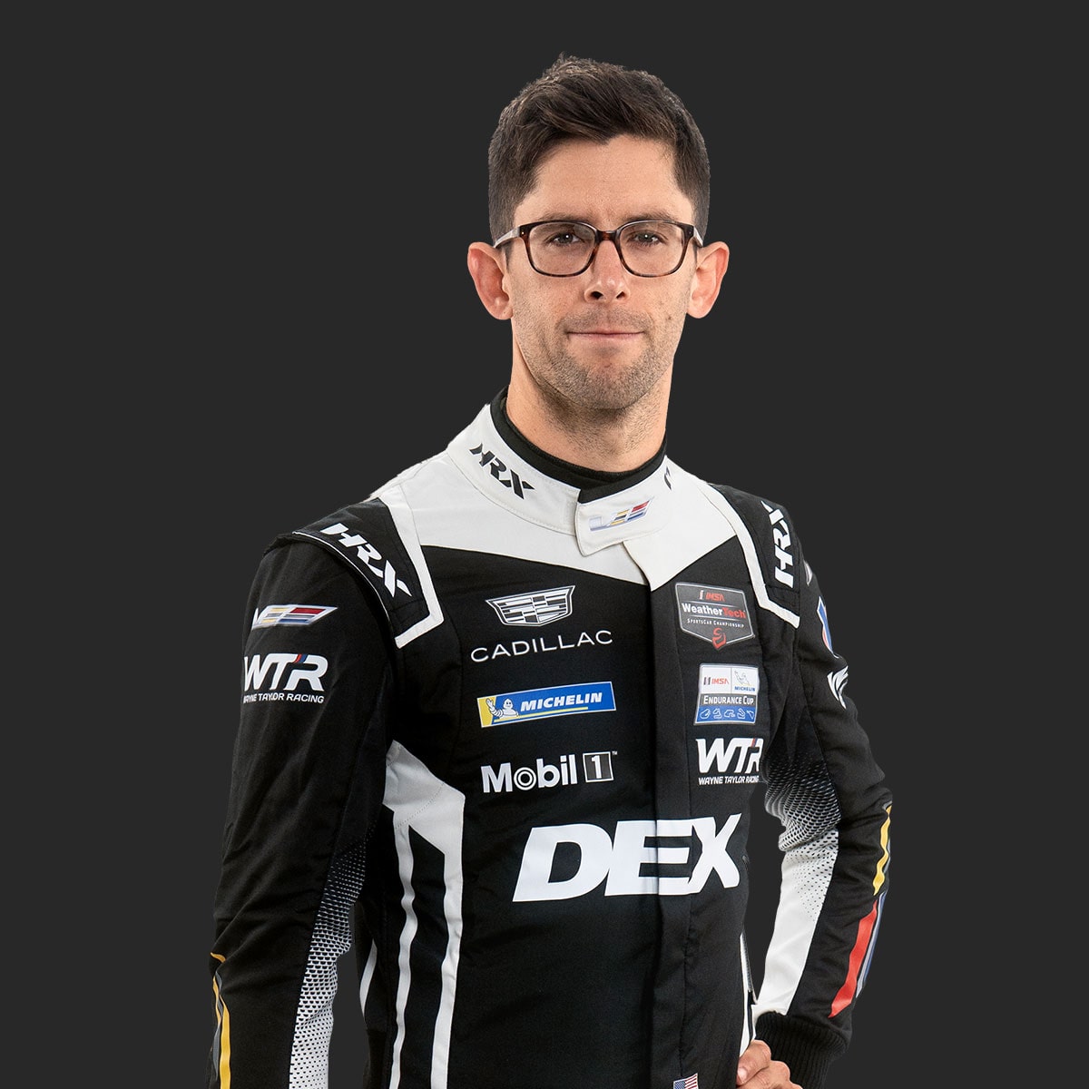 Cadillac Racing Team Driver Jordan Taylor Wearing a Black and White DEX Imaging–Branded Racing Suit With Sponsor Patches Stands Against a Solid Dark Background.
