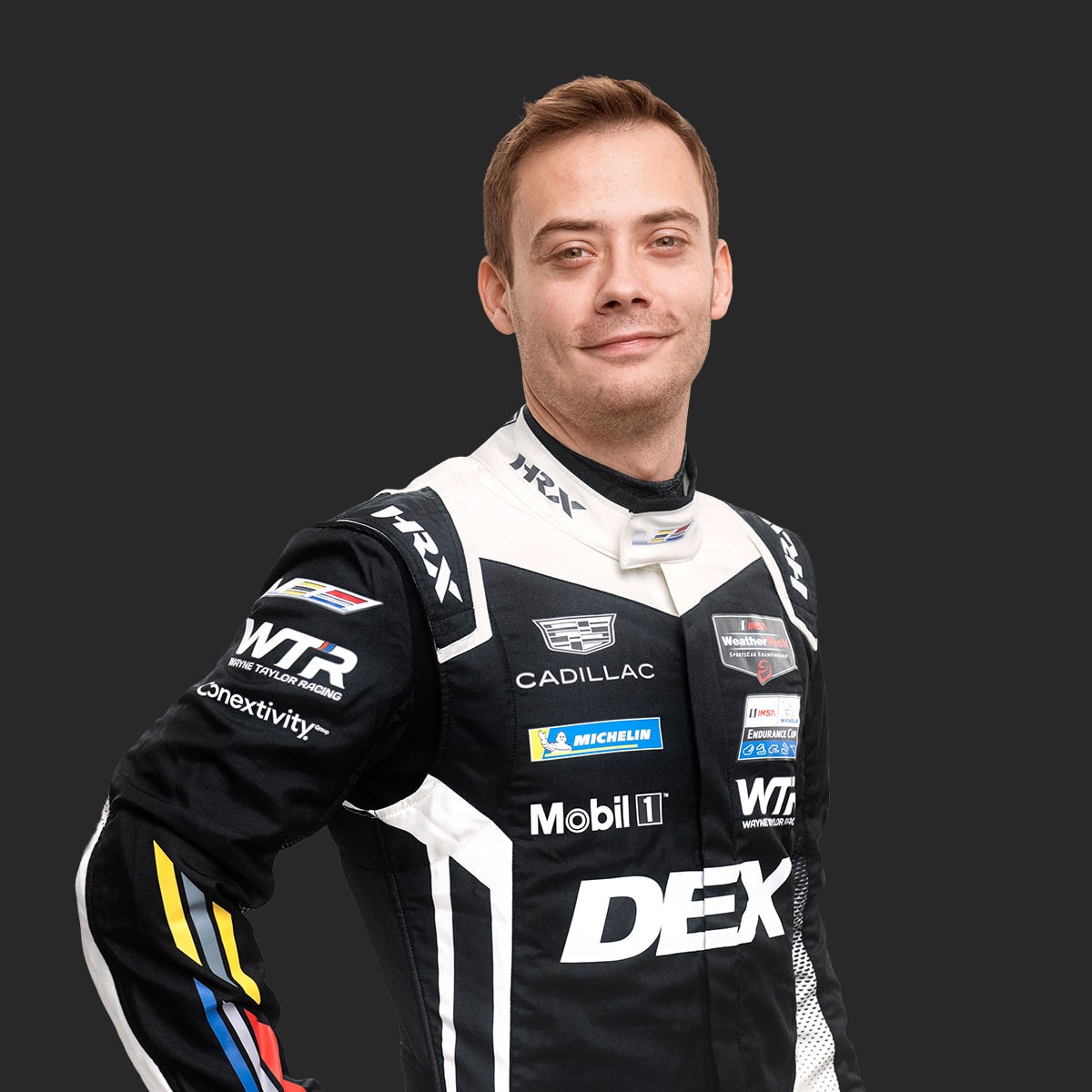 Cadillac Racing Team Driver Louis Deletraz Wearing a Black and White DEX Imaging–Branded Racing Suit With Color Accents on the Sleeves Stands With One Arm Bent Against a Solid Dark Background.