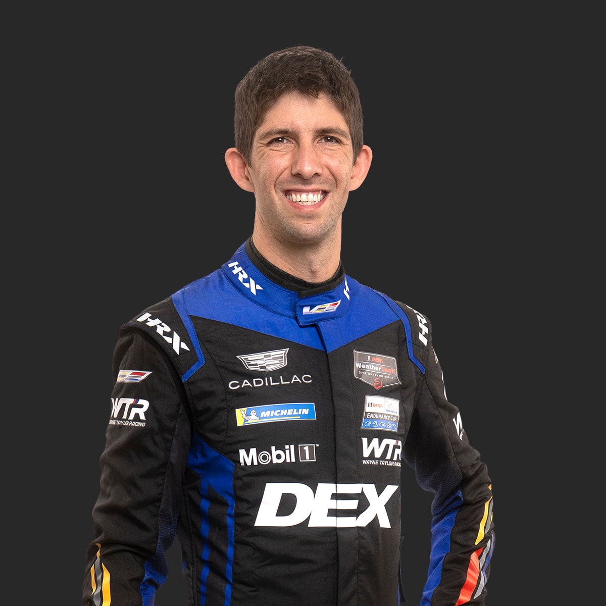 Cadillac Racing Team Driver Ricky Taylor Wearing a Blue and Black DEX Imaging–Branded Racing Suit Is Shown Standing Against a Solid Dark Background.