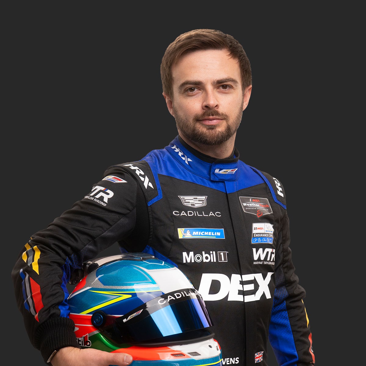 Cadillac Racing Team Driver Will Stevens Wearing a Blue and Black DEX Imaging–Branded Racing Suit and Holding a Multicolored Racing Helmet Is Shown Standing Against a Solid Dark Background.